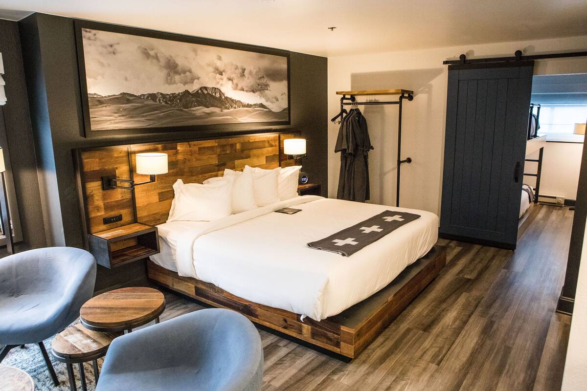 Ein modernes Hotelzimmer mit einem Holzkopfteil, einem großen Bett, zwei Nachttischen, einem an der Wand befestigten Fernseher und einem Gemälde von einer Berglandschaft.