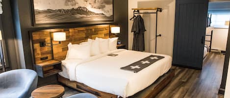 A modern hotel room with a wooden headboard, a large bed, two bedside tables, a wall-mounted TV, and a painting of a mountain landscape.