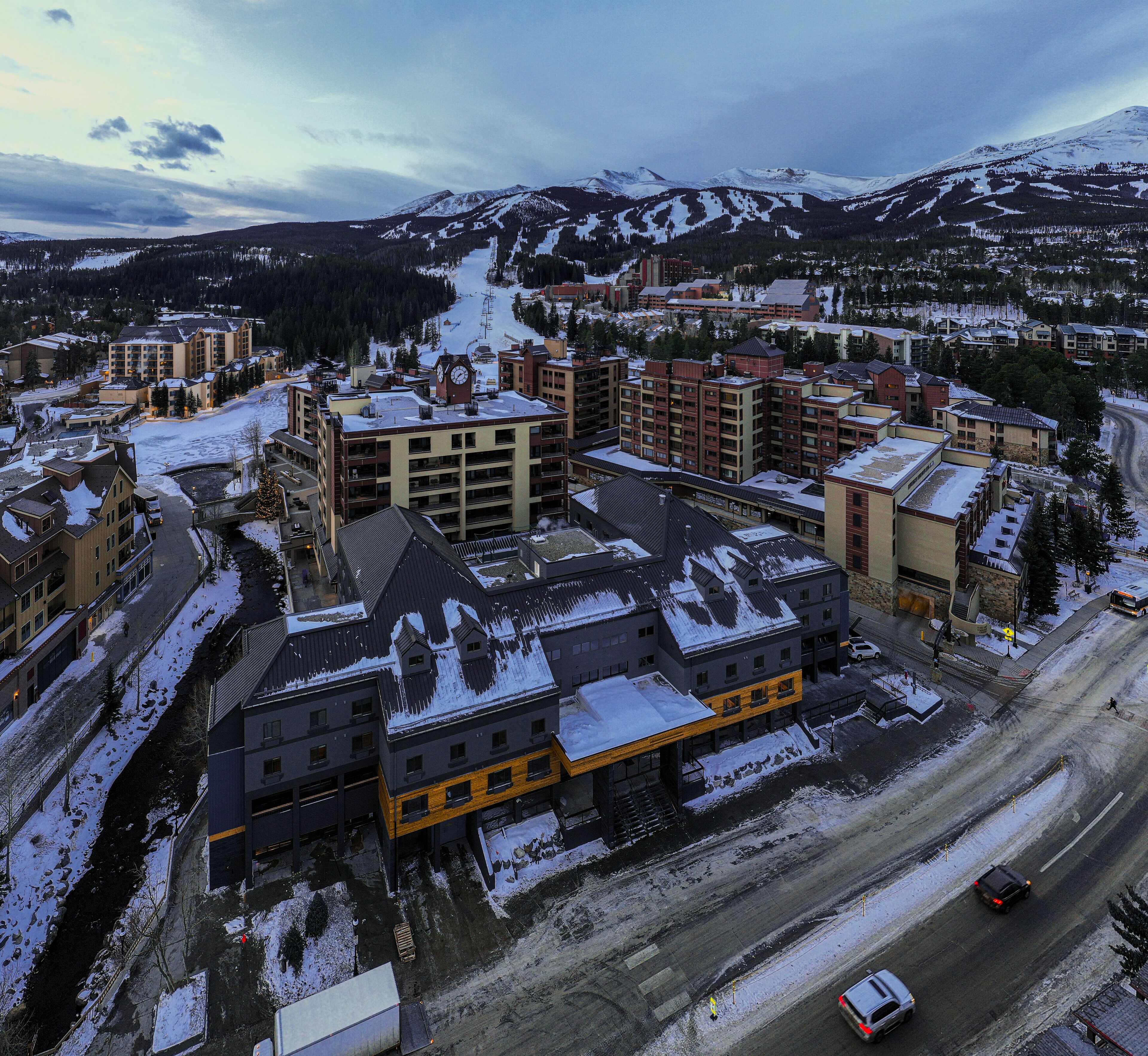 Un hotel de varios pisos con una torre del reloj, rodeado de edificios cubiertos de nieve y un paisaje montañoso.
