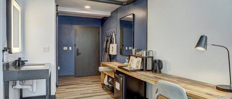 A modern hotel room with a wooden desk, a black refrigerator, a sink, a mirror, and a lamp.