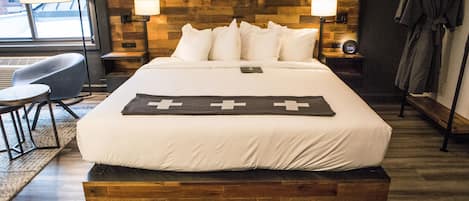 Una habitación de hotel moderna con una cama grande, cabecera de madera, dos lámparas de noche, una mesita de noche, una silla y vistas al exterior.