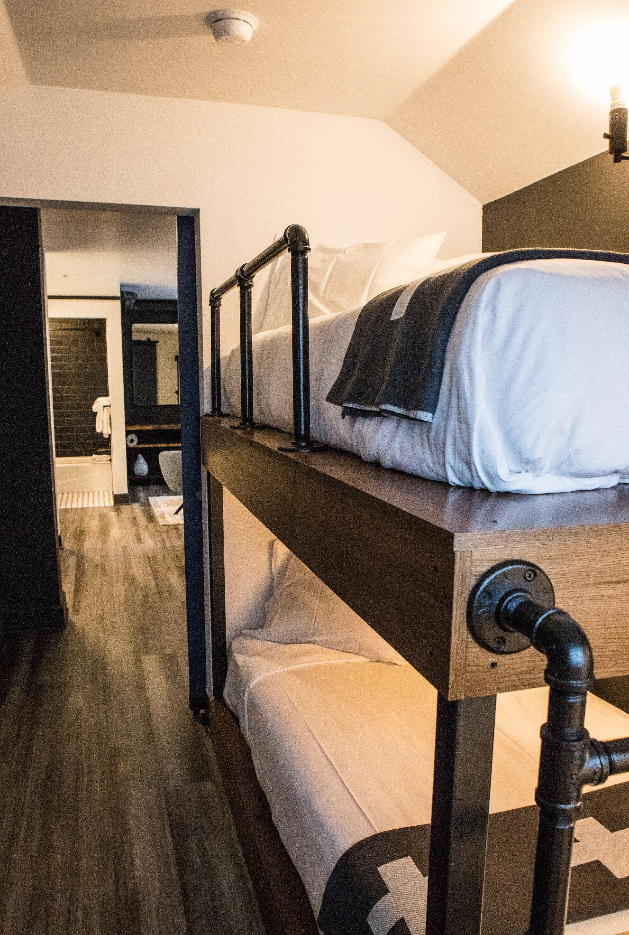 Una cama individual con estructura de madera y tuberías de metal, un baño con ducha e inodoro, y un espejo.