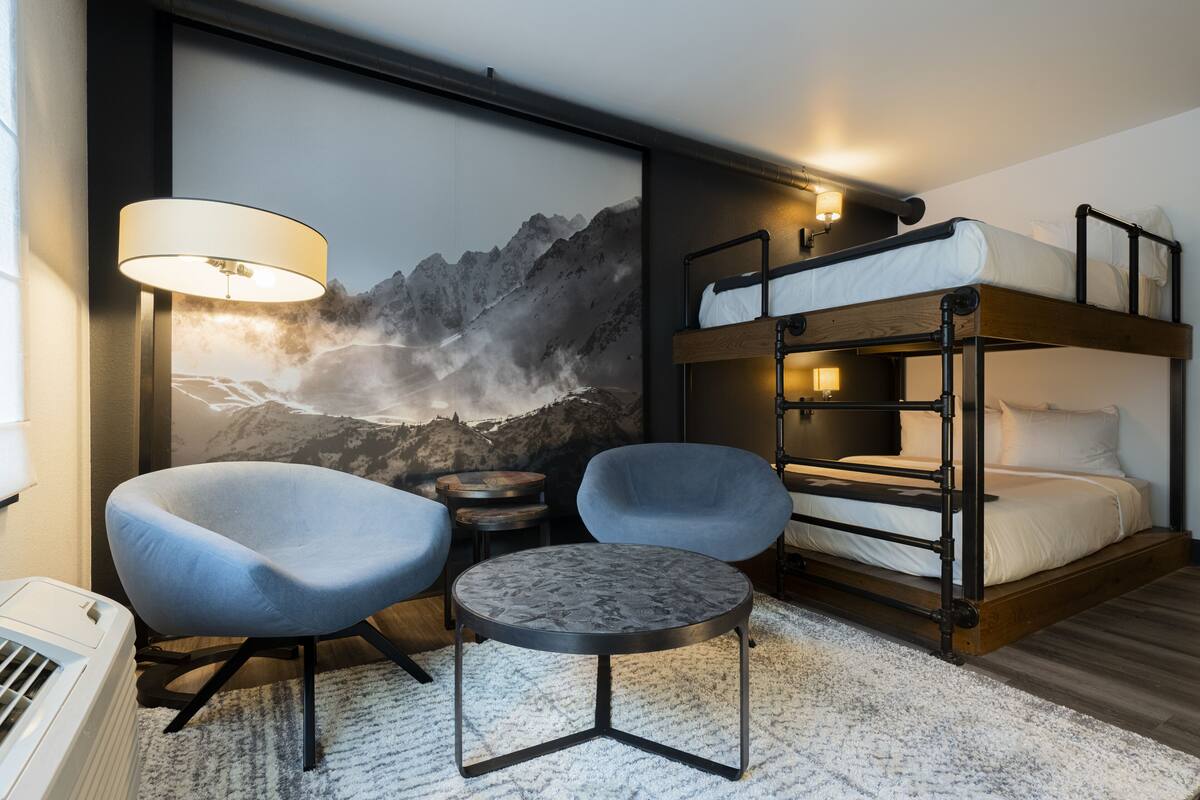 Ein modernes Hotelzimmer mit einem Stockbett, zwei Sesseln, einem runden Tisch und einer großen Wandmalerei mit Bergblick.