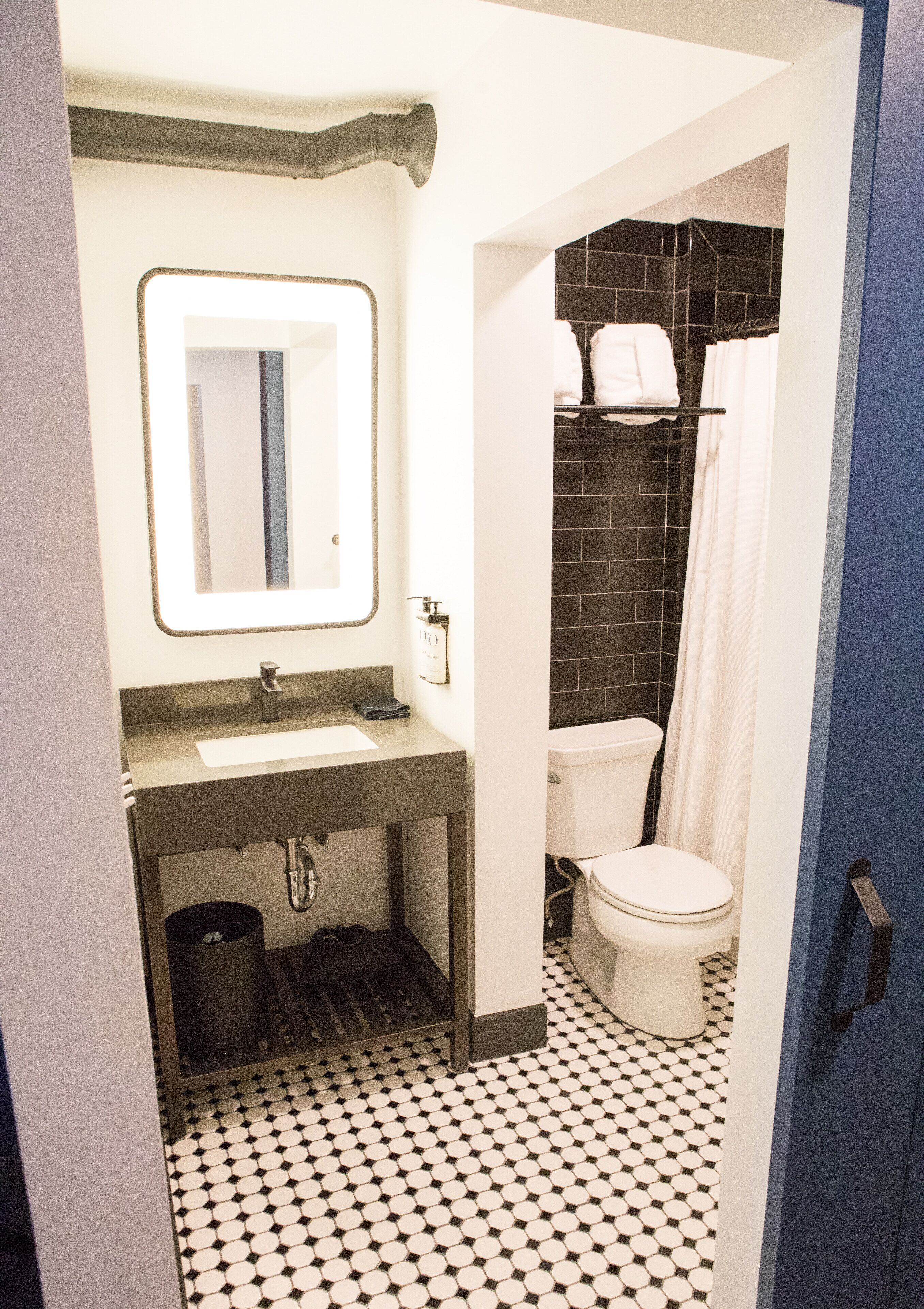 Baño con lavamanos, espejo y retrete blancos, e igualmente una zona de ducha con azulejos negros.