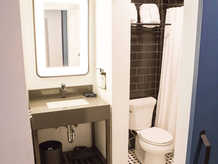 Baño con lavamanos, espejo y retrete blancos, e igualmente una zona de ducha con azulejos negros.