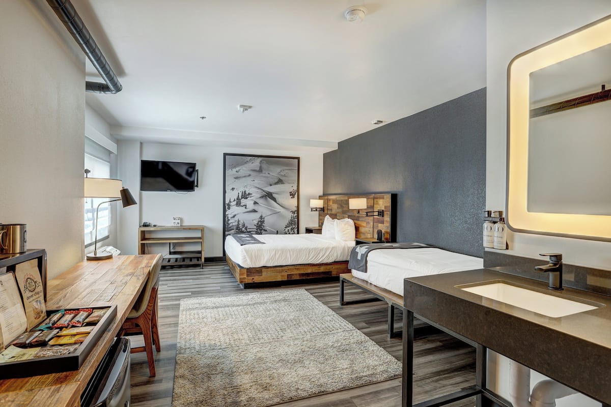 Ein modernes Hotelzimmer mit einem Holztisch, einem großen Bett, einem Nachttisch, einem Badezimmer mit Spiegel und einem an der Wand montierten Fernseher.