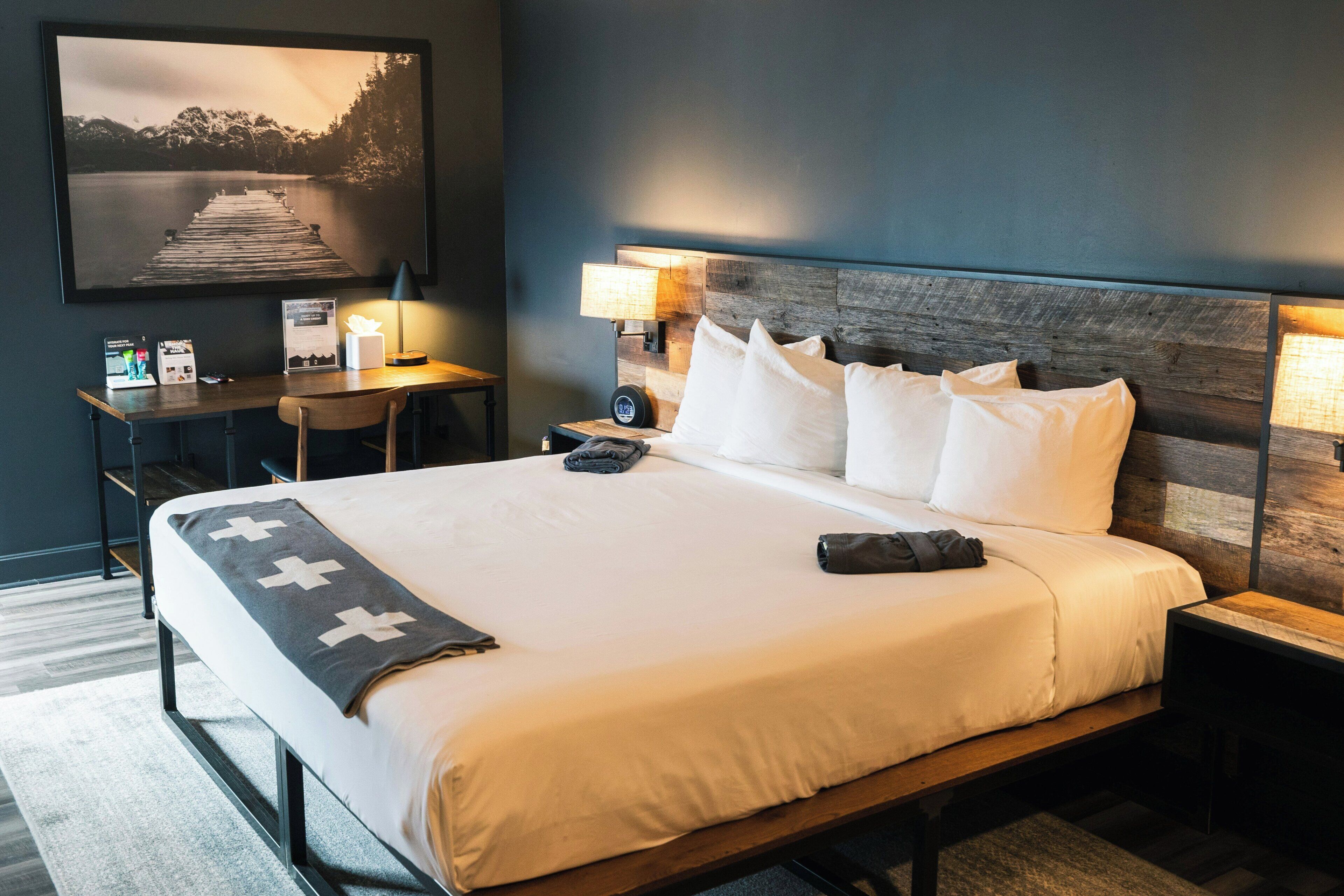 A neatly made bed with white linens, a wooden headboard, and a desk with a lamp.