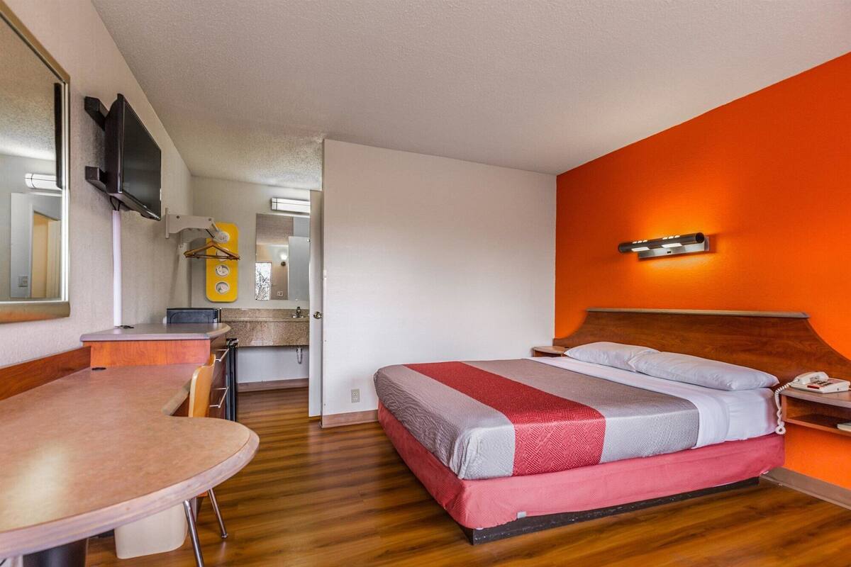 A hotel room with a wooden floor, a bed with a red and grey bedspread, a wooden headboard, a bedside table with a phone, and a mirror on the wall.