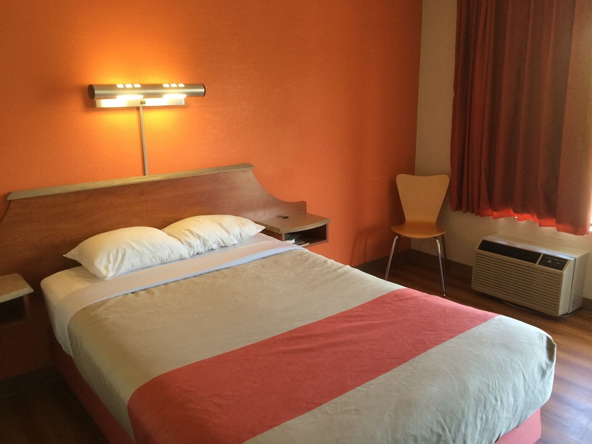 A hotel room with a bed, a nightstand, a chair, a wall-mounted lamp, and an air conditioning unit.