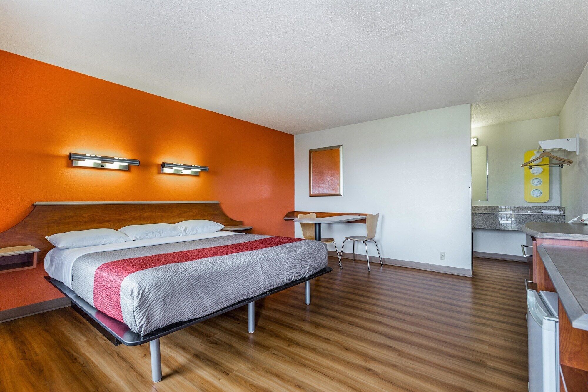 Une chambre d’hôtel avec un plancher en bois, un lit recouvert d’une couverture rouge et blanche, un bureau avec une chaise, et une salle de bain équipée d’un miroir et d’un porte-serviettes.