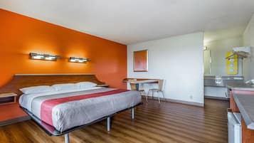 Quarto de hotel com piso de madeira, uma cama com manta vermelha e branca, uma escrivaninha com cadeira e um banheiro com espelho e porta-toalhas.