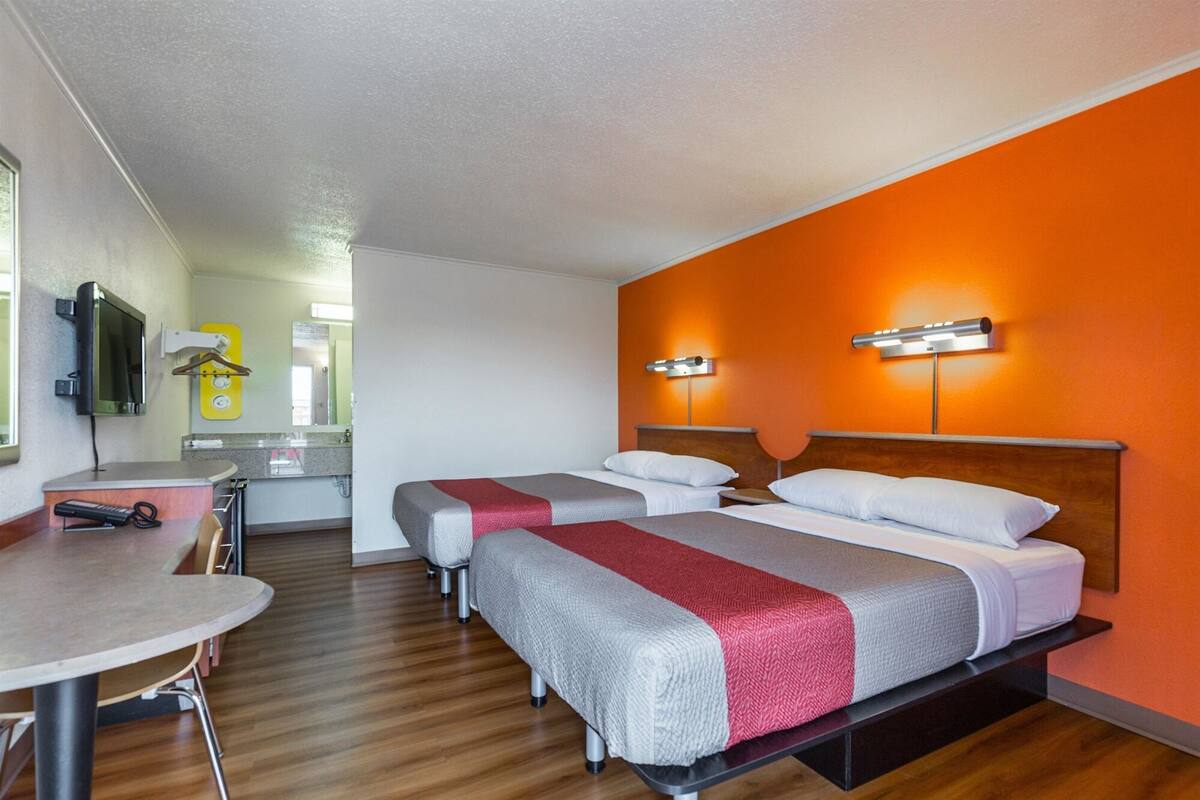 A hotel room with two beds, a small table, a TV, and a bathroom visible in the background.