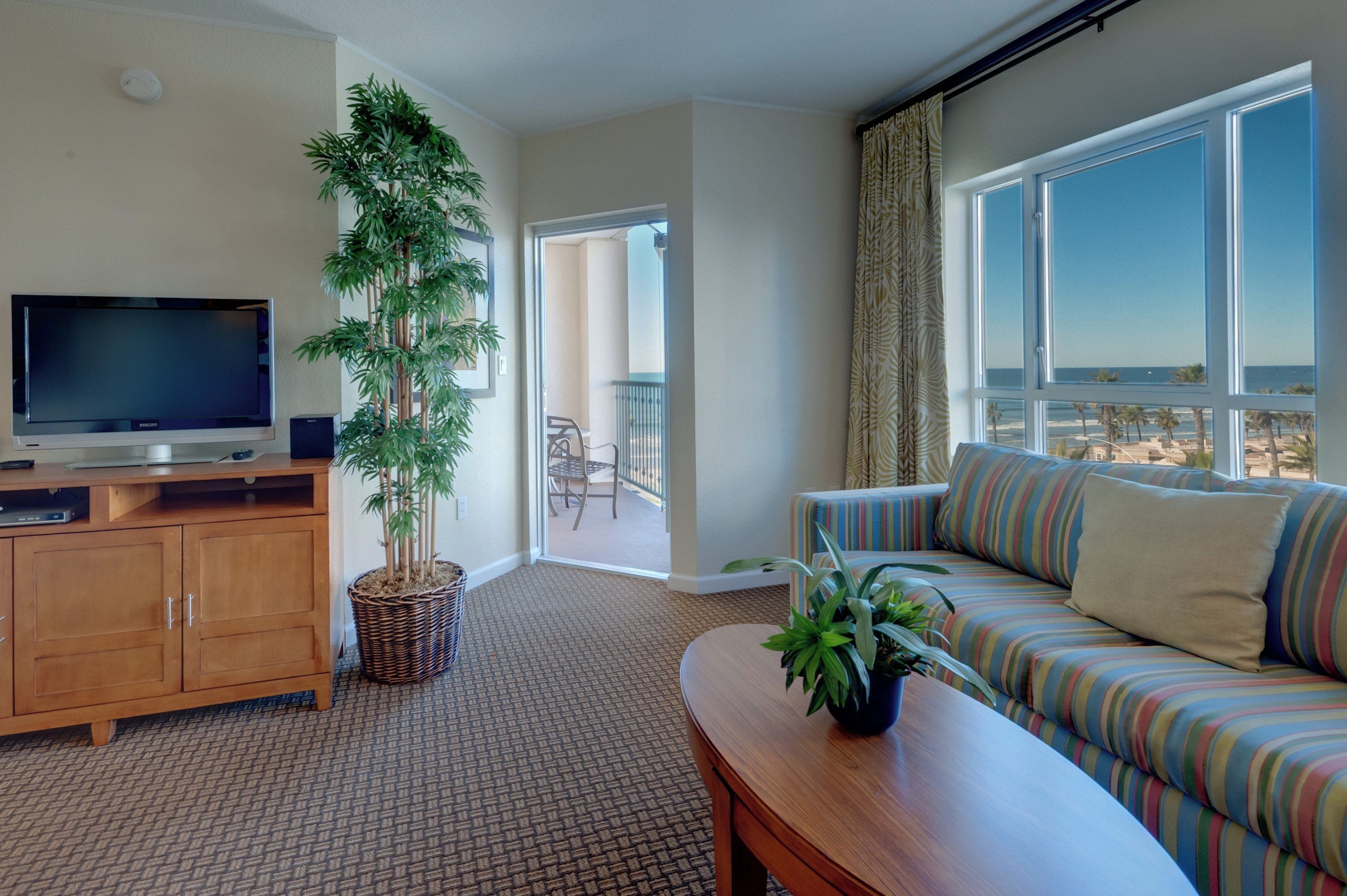 Habitación de hotel con televisión, sofá, mesa de comedor, planta en maceta y vista a la playa.