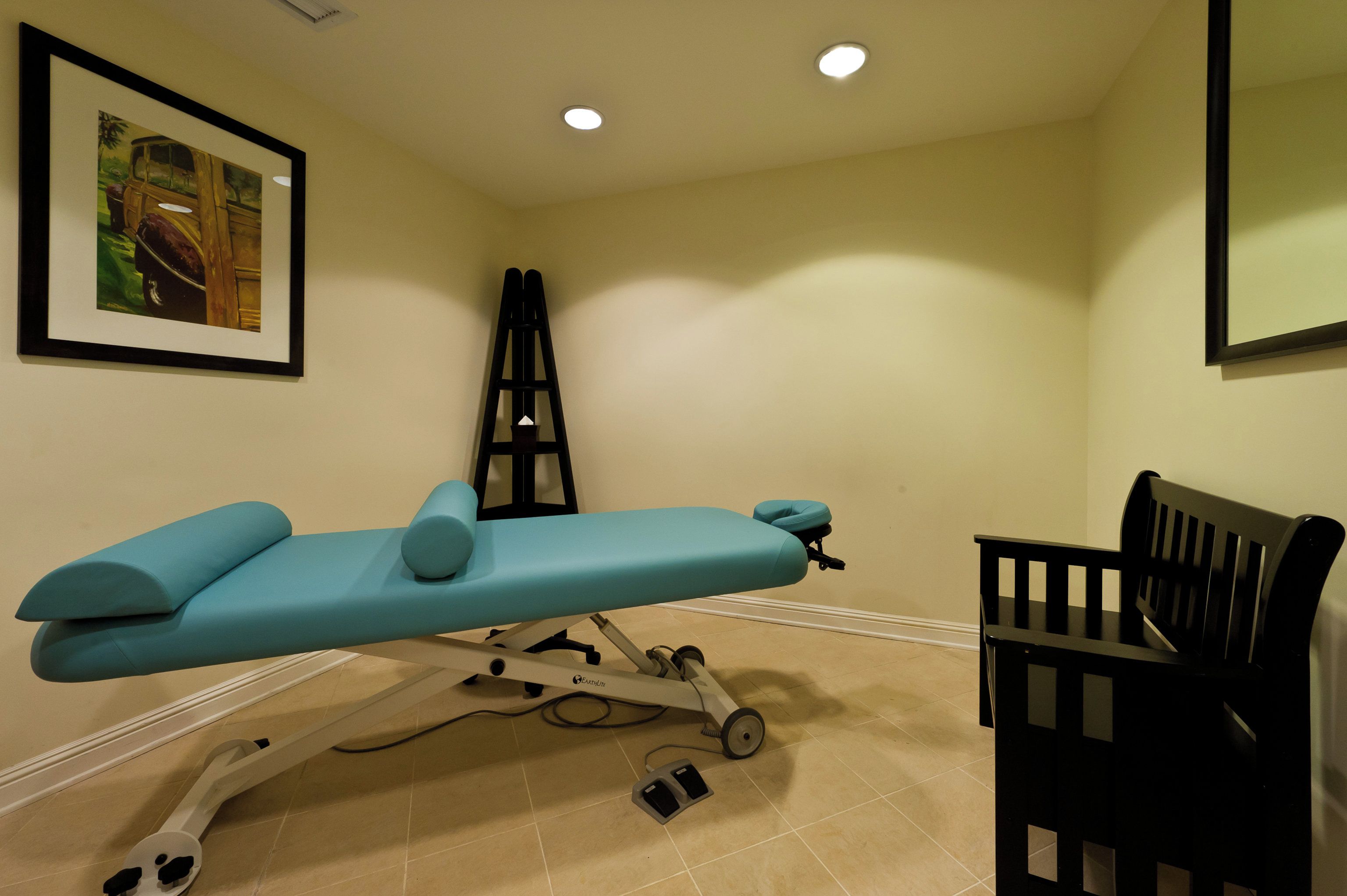 Une table d’examen médical sur roulettes, une chaise noire et un tableau encadré accroché au mur.