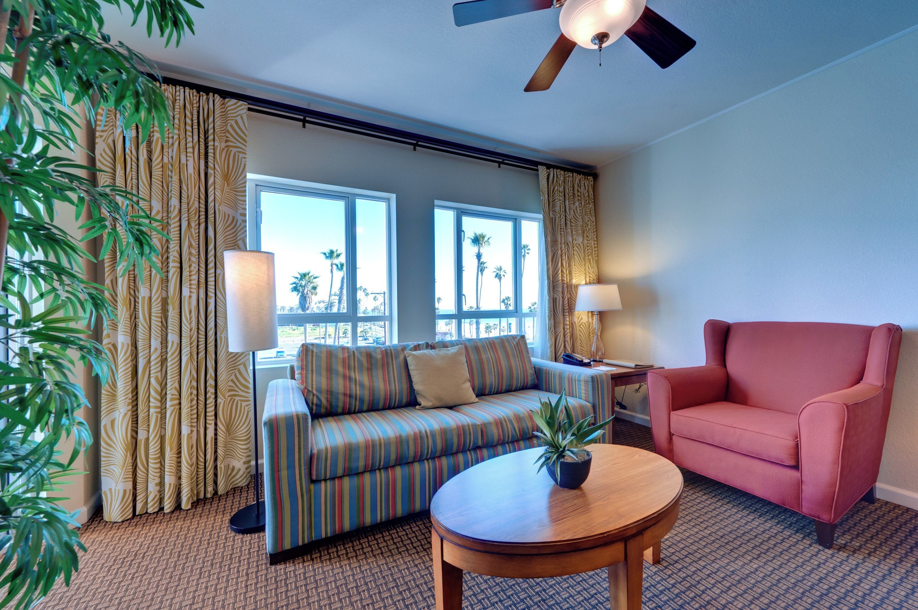 A hotel room with a striped sofa, a pink armchair, a round wooden table, and a ceiling fan.