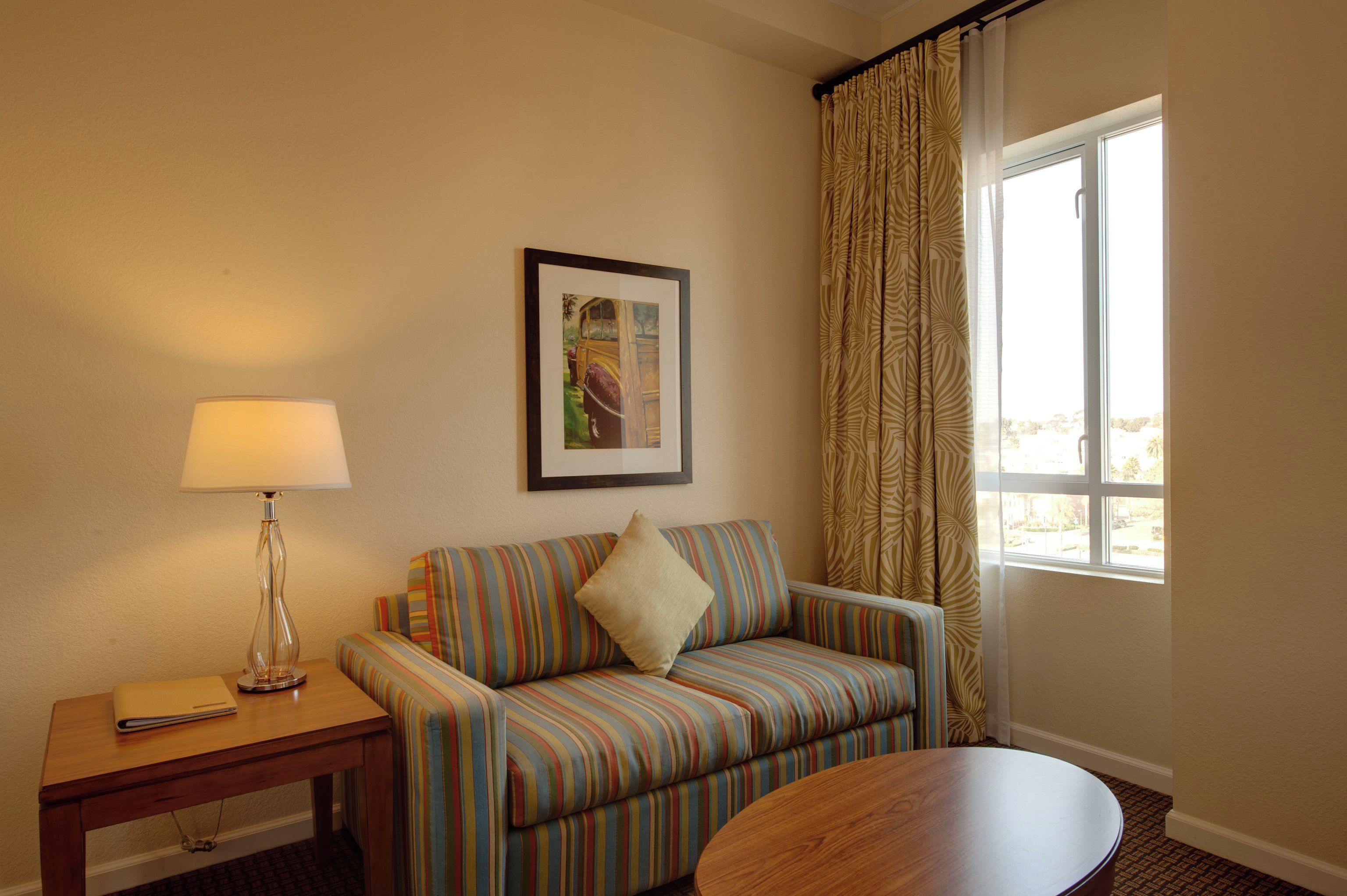 Une chambre d’hôtel avec un canapé à rayures, une petite table avec une lampe, un tableau encadré au mur et une fenêtre avec des rideaux.