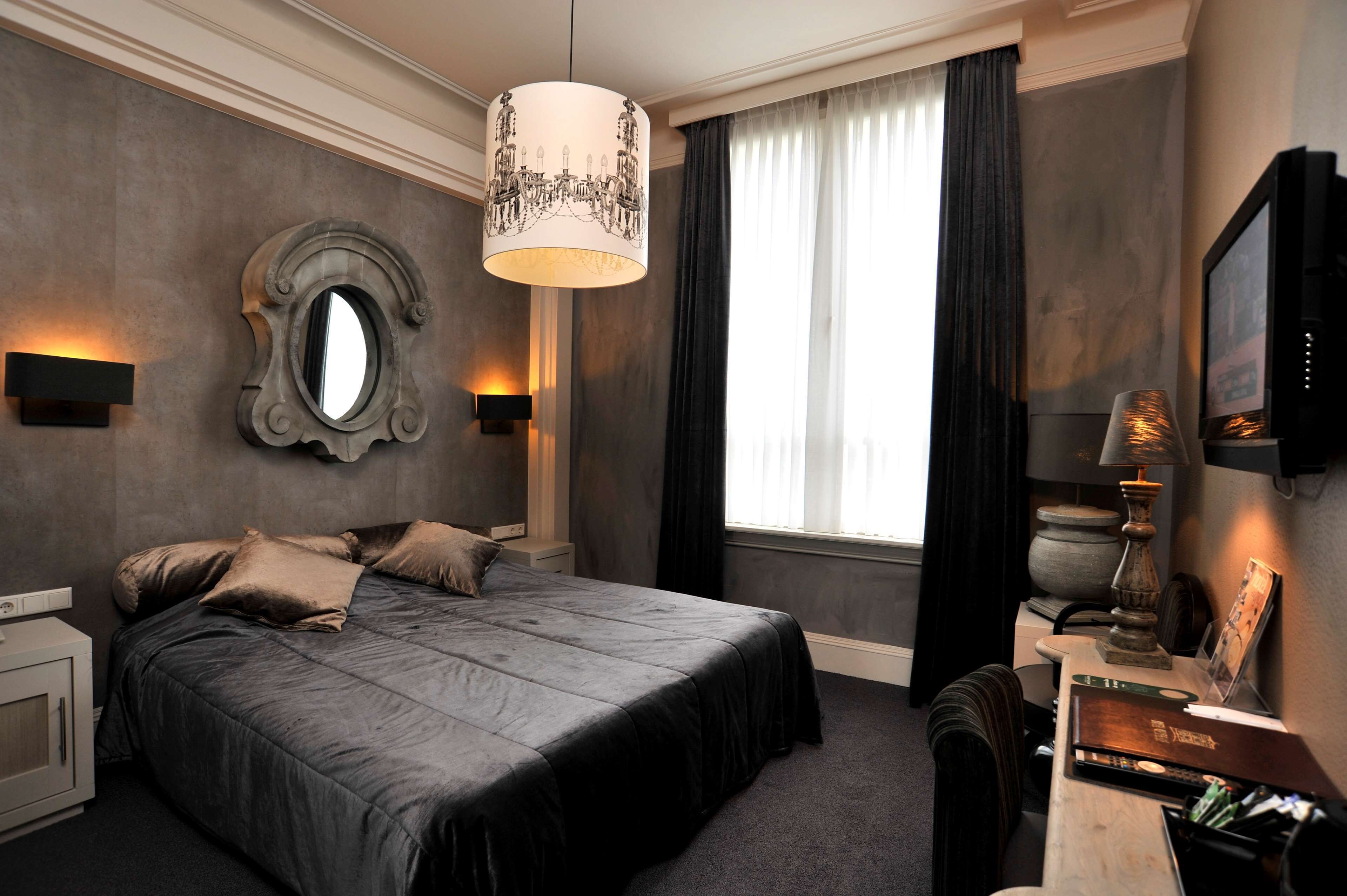A bedroom with a large bed, a round mirror, two wall-mounted lamps, a desk with a lamp, and a television.