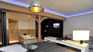A modern hotel room with a large bed, a desk, a TV, a sofa, and a bathtub. - Golden Tulip West Ende (Helmond)