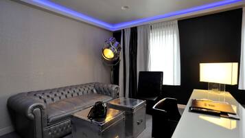 Une chambre d’hôtel moderne avec un canapé en cuir gris, une table basse en verre, deux valises métalliques, un bureau avec une lampe et un téléviseur à écran plat.