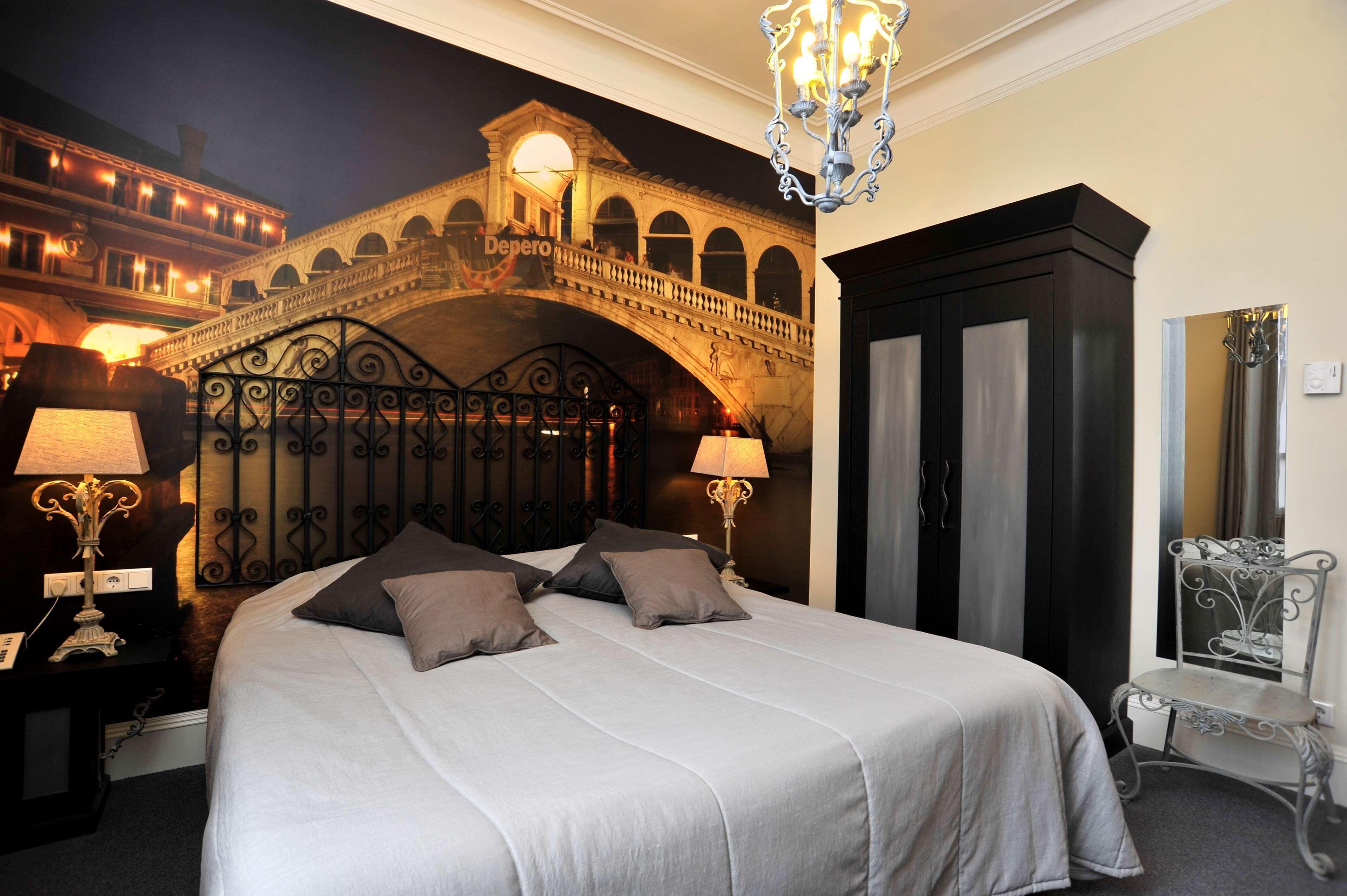 Una habitación de hotel con una cama, dos lámparas de noche, un armario negro, un espejo y una araña.