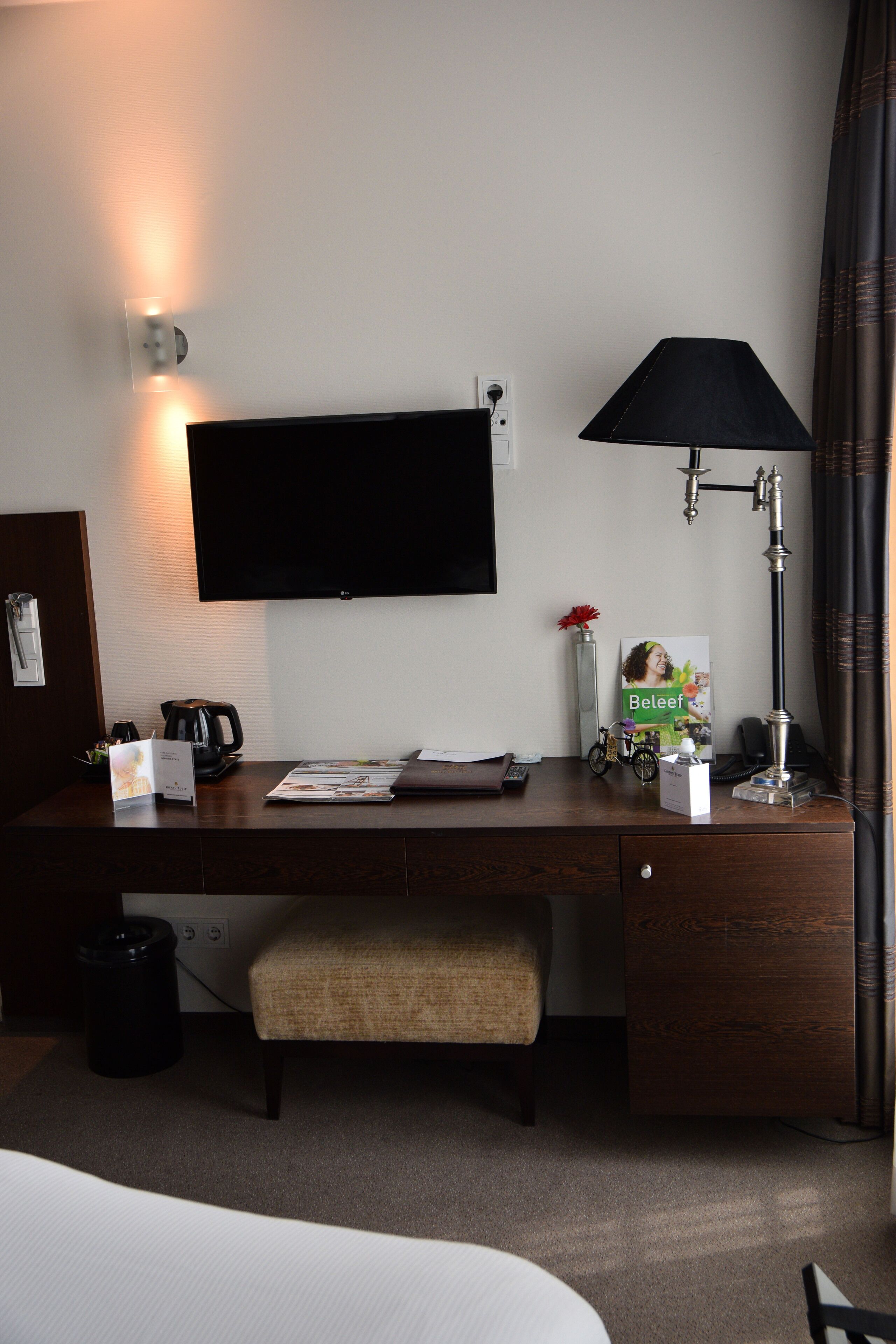 Habitación de hotel con cama, escritorio con lámpara, televisor, cafetera y una planta pequeña.