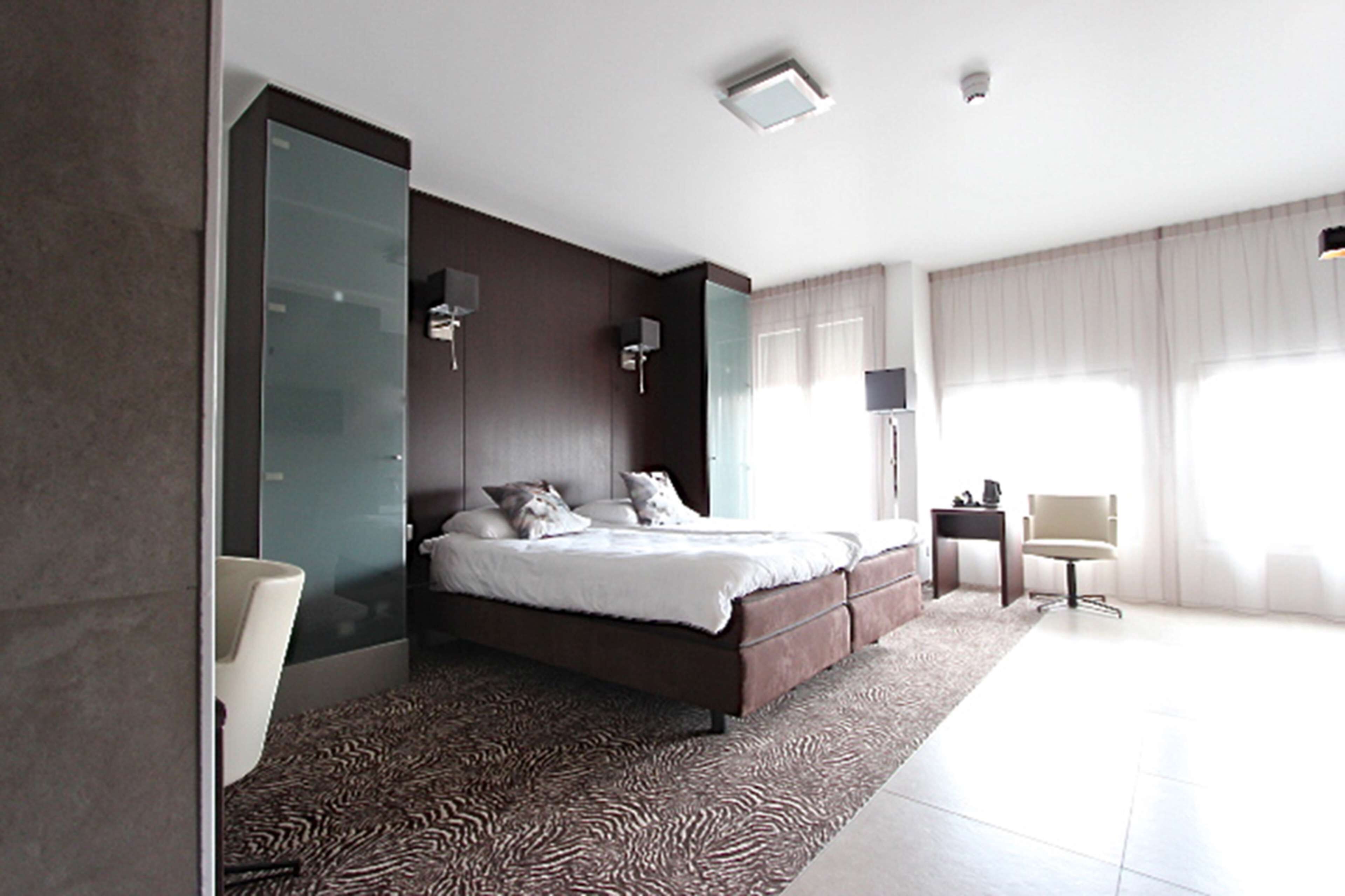 A modern bedroom with a large bed, a glass partition wall, a small bedside table, and a chair.