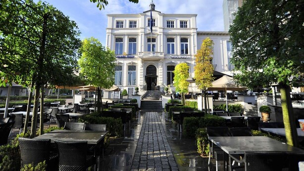 A building with a central entrance, surrounded by outdoor seating areas with tables and chairs. - Golden Tulip West Ende (Helmond)