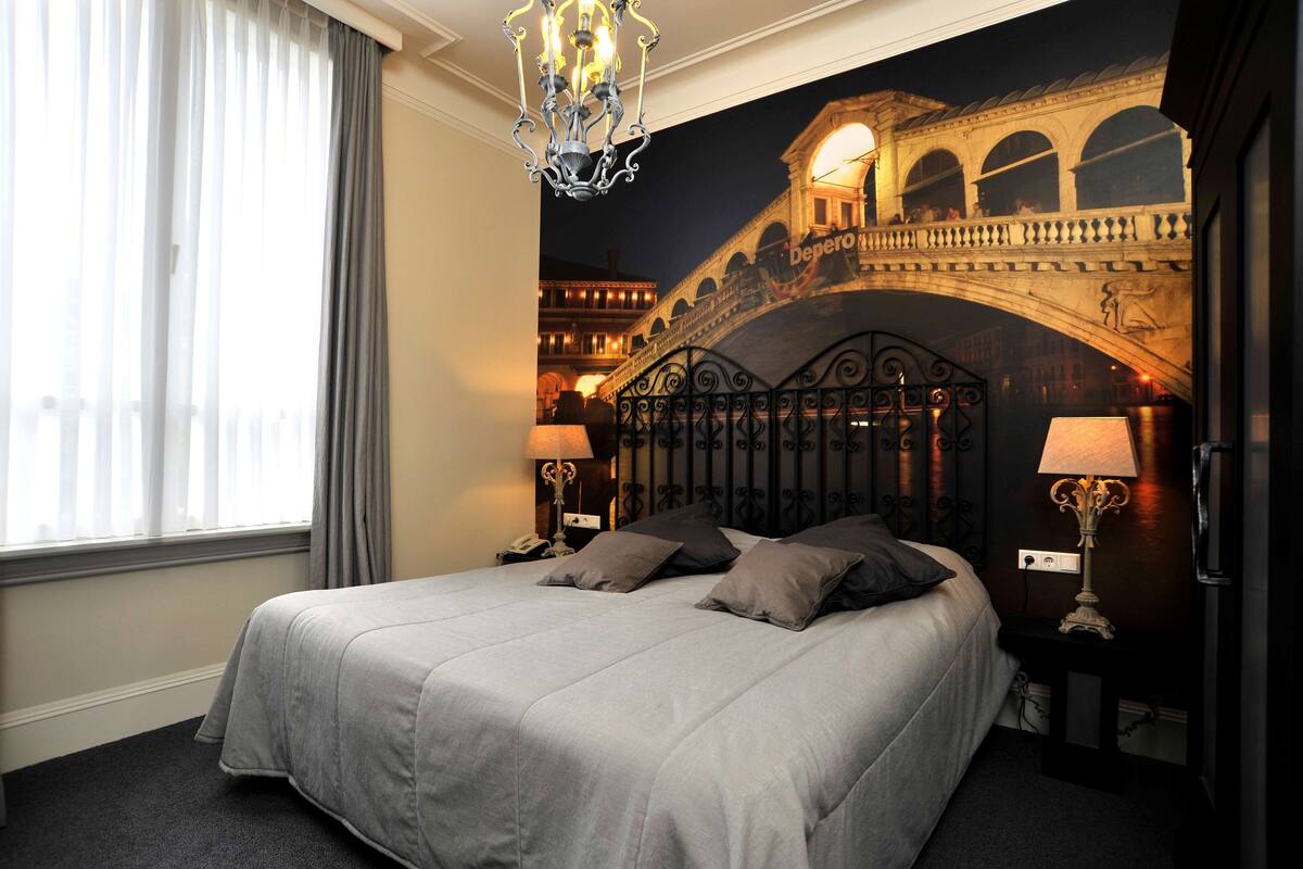 Ein Hotelzimmer mit einem Bett, zwei Nachttischen mit Lampen, einem Fenster mit Vorhängen und einem Wandbild einer Brücke.