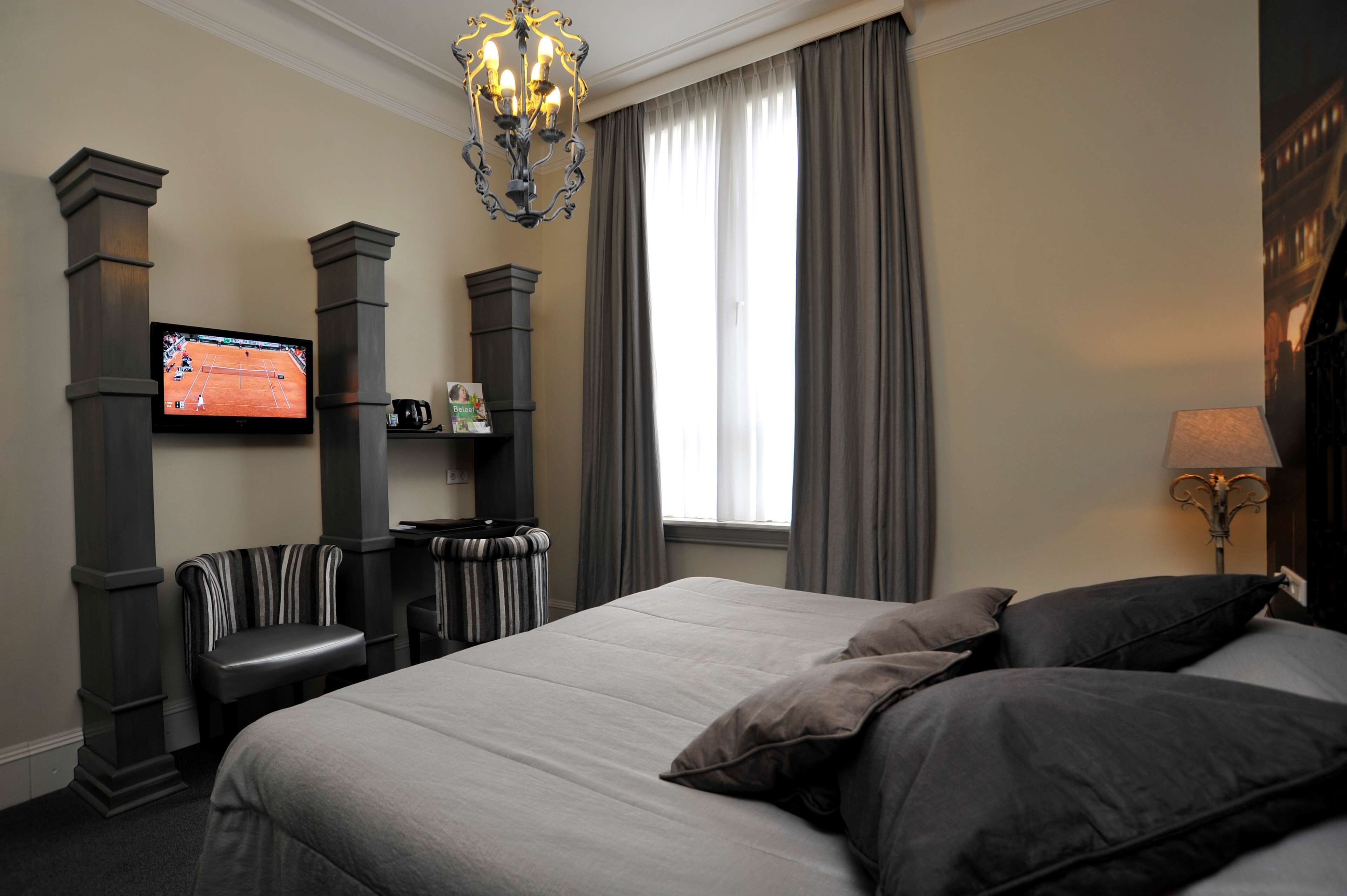 A hotel room with a large bed, a television mounted on a pillar, a bench, a small table with a magazine, and a window with curtains.