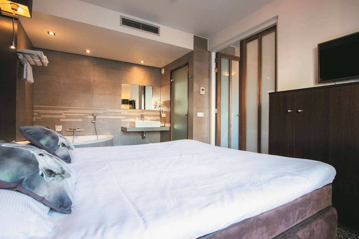 Ein modernes Hotelzimmer mit einem großen Bett, einem Badezimmer mit Waschbecken und Spiegel sowie einem an der Wand befestigten Fernseher.