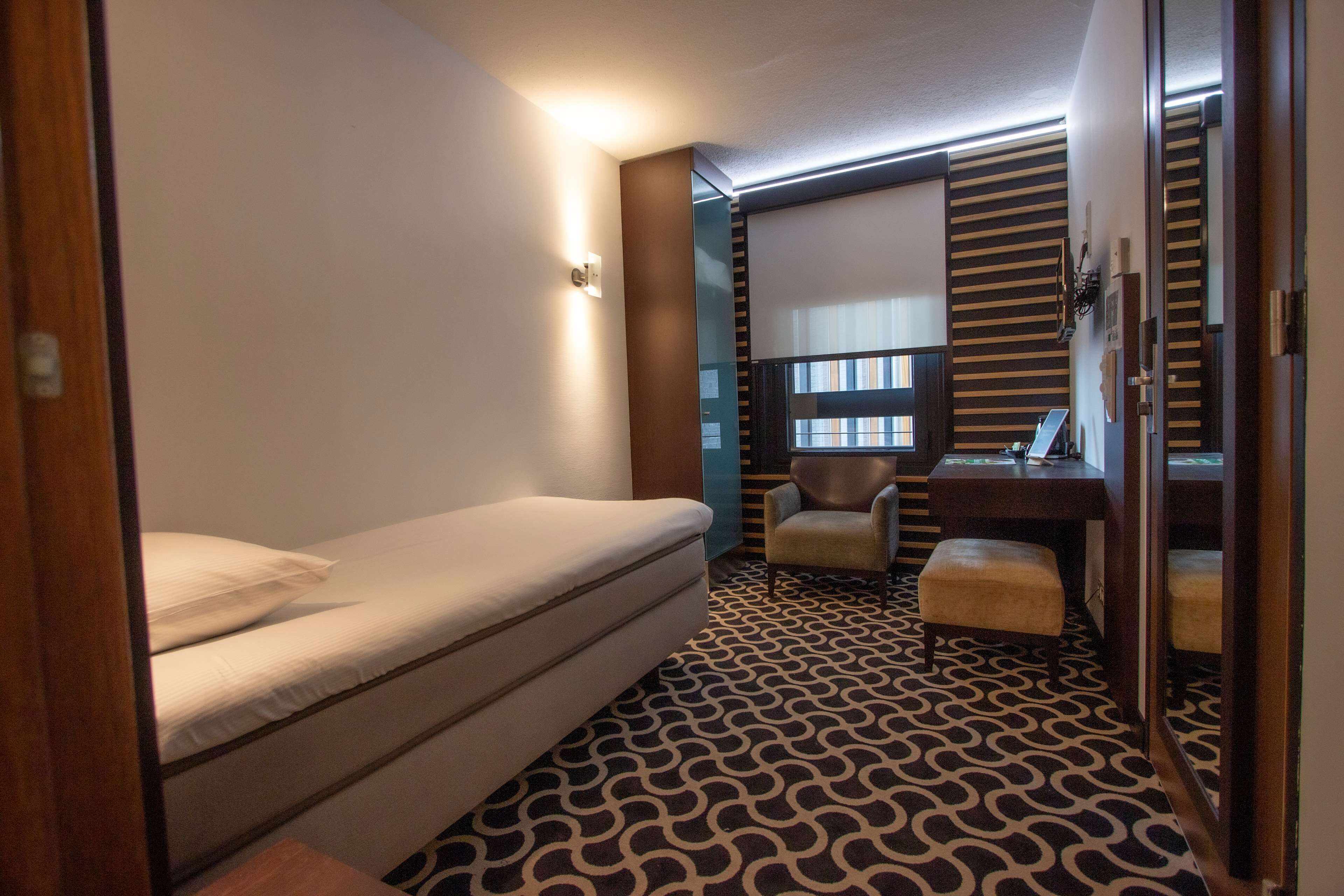 A compact room with a bed, a desk, a chair, and a patterned floor.