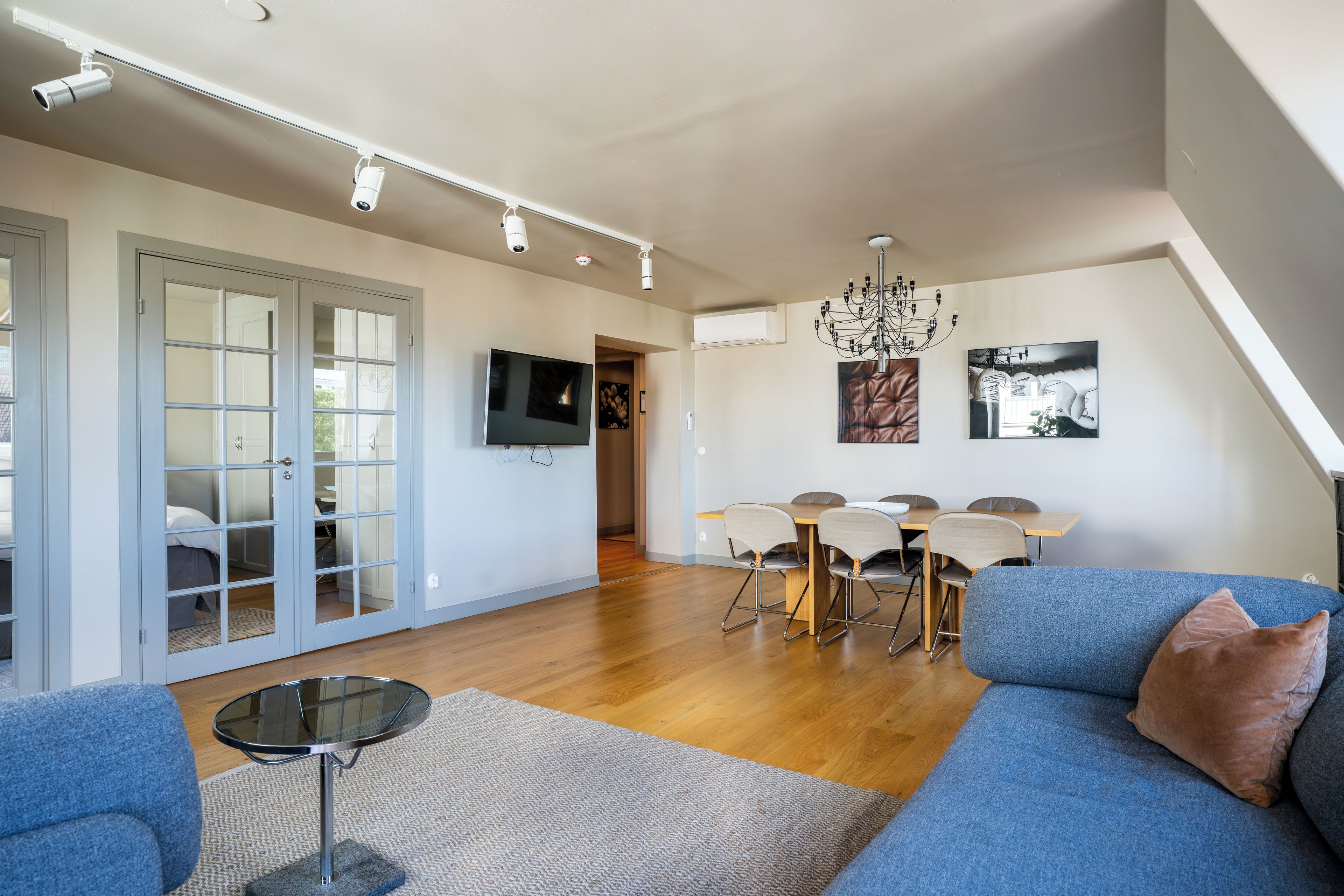 A modern living room with a blue sofa, a round glass table, and a dining area with a wooden table and chairs.