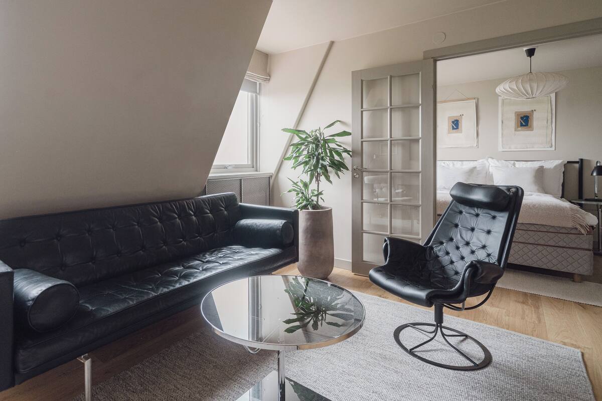 A modern living room with a black leather sofa, a glass coffee table, a black leather chair, and a large potted plant.