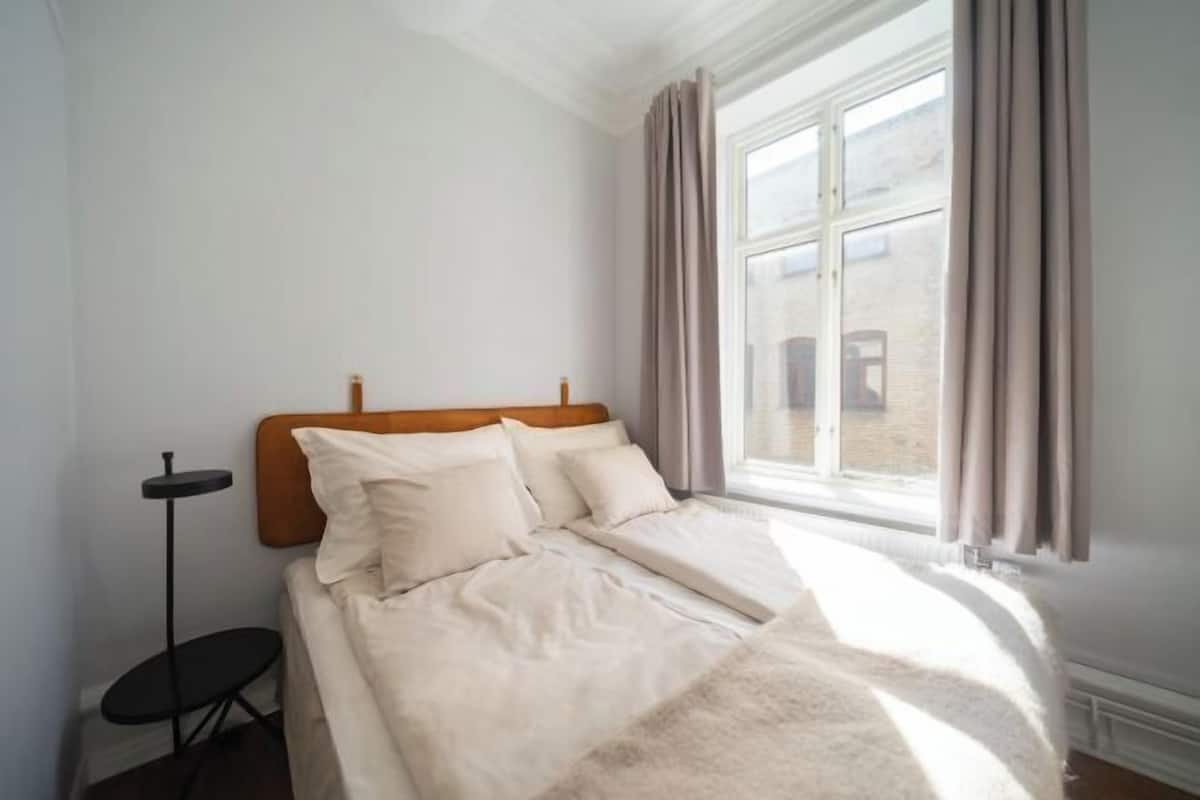 A bedroom with a bed, a small table, and a window with curtains.