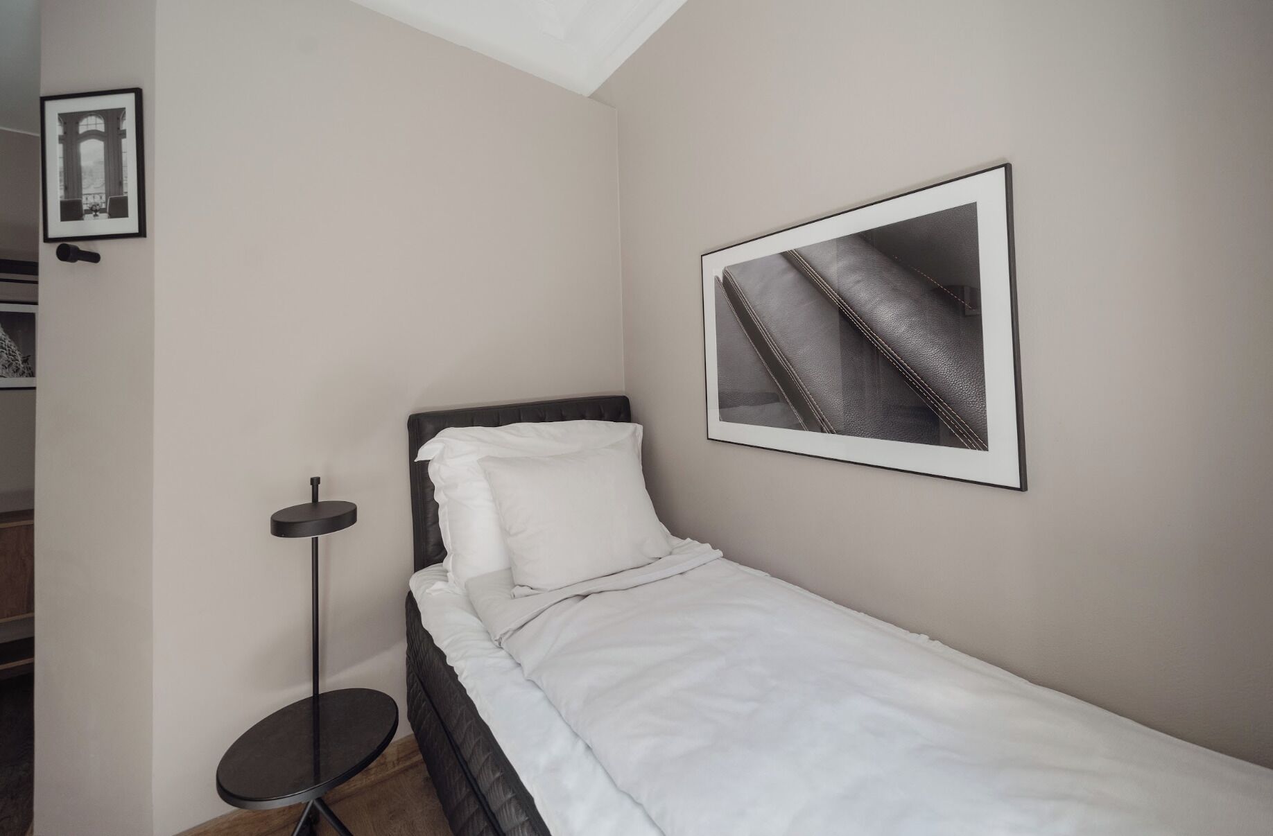 Ein kleines, ordentlich bezogenes Bett mit weißer Bettwäsche in einem Zimmer mit einem runden Tisch und einem gerahmten Bild an der Wand.