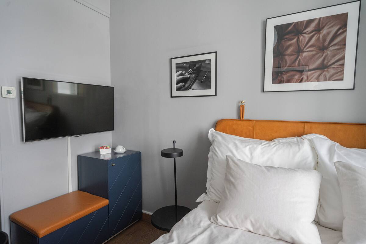A hotel room with a bed, a TV, a small cabinet, a round floor lamp, and two framed pictures on the wall.