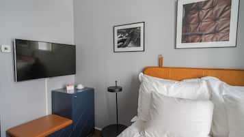 A hotel room with a bed, a TV, a small cabinet, a round floor lamp, and two framed pictures on the wall.