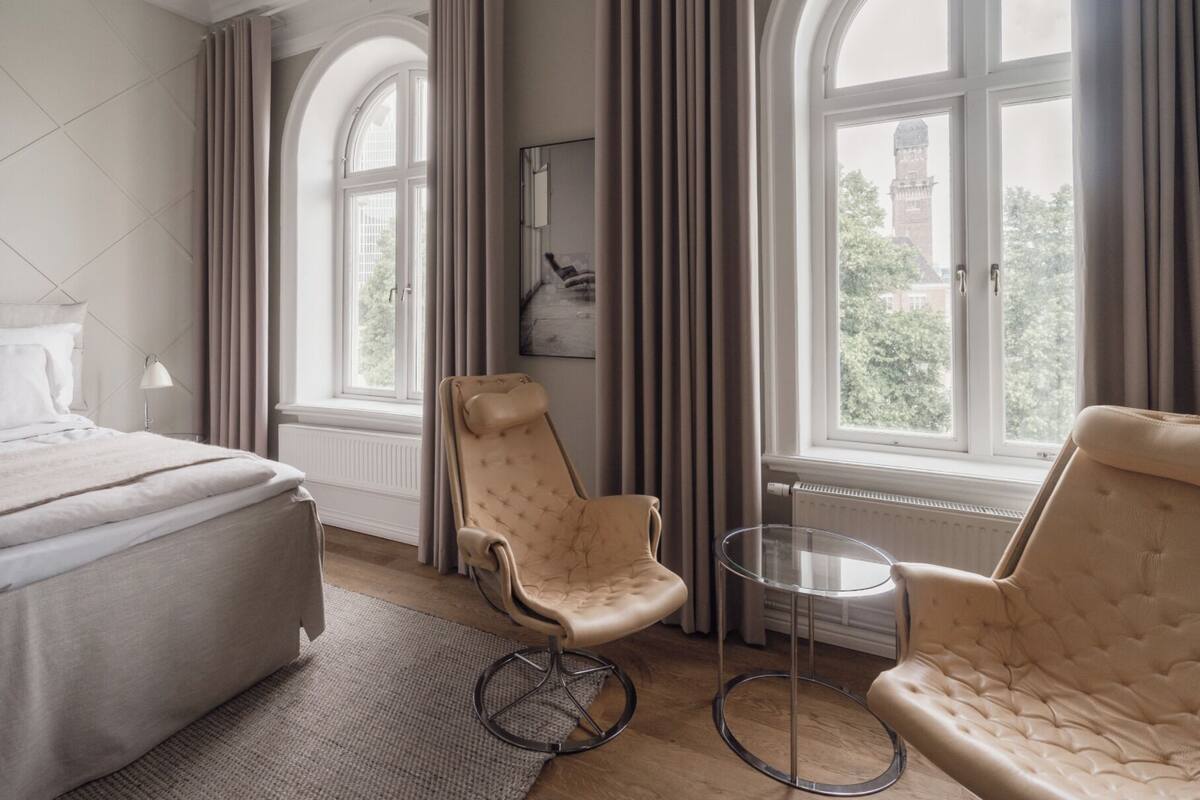 A hotel room with a bed, two armchairs, a small table, and large windows with curtains.