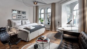 Une chambre moderne avec un grand lit, un coin salon comprenant un canapé et un fauteuil, une table basse ronde en verre, et une vue sur la ville grâce à de grandes fenêtres.