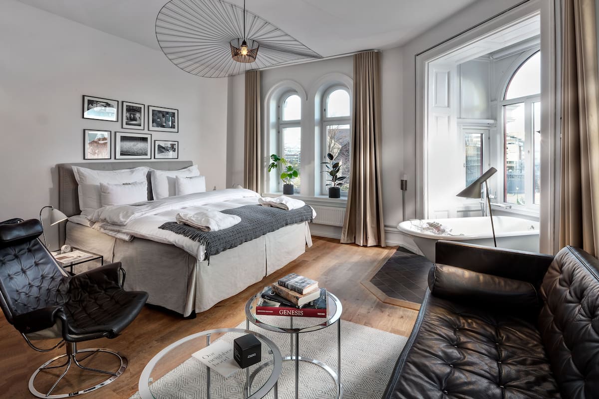A modern bedroom with a large bed, a sitting area with a sofa and armchair, a round glass coffee table, and a view of the city through large windows.