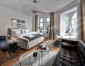 Un dormitorio moderno con una cama grande, una zona de estar con un sofá y un sillón, una mesa de centro redonda de cristal y vistas a la ciudad a través de amplias ventanas.