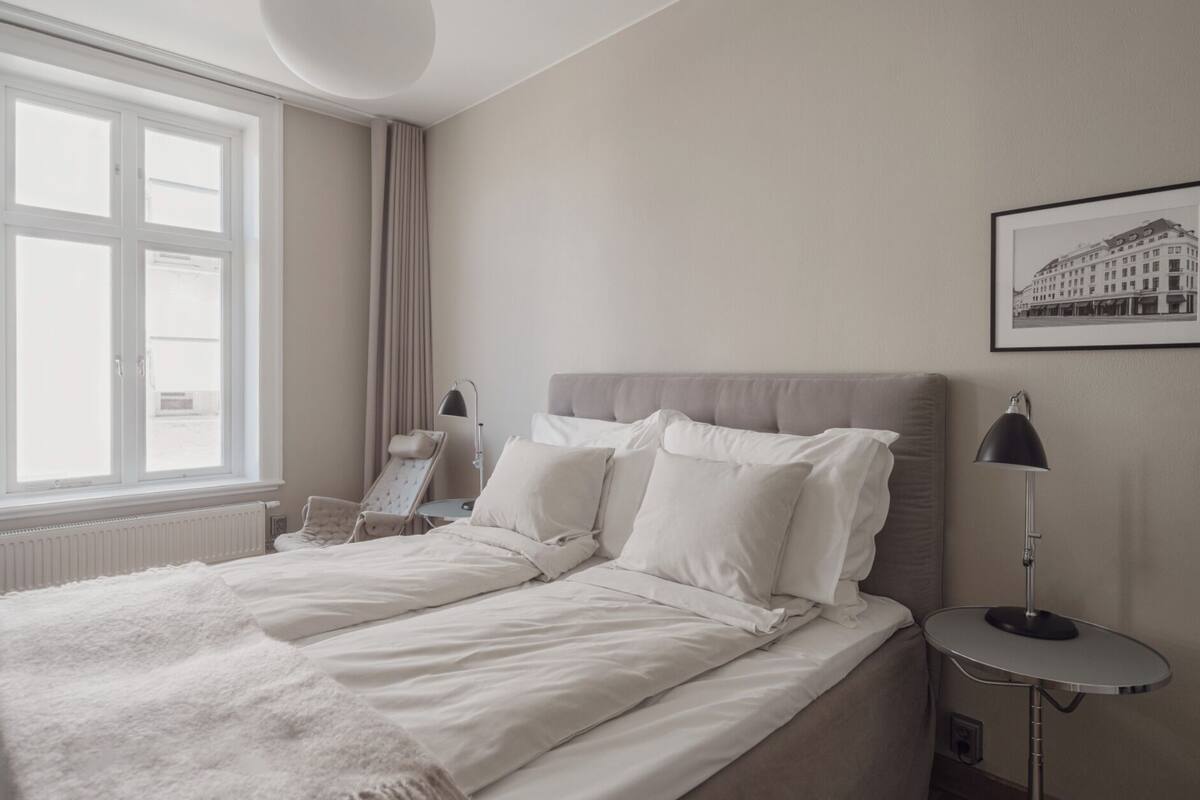 A bedroom with a bed, a bedside table, a chair, a window with curtains, and a framed picture on the wall.