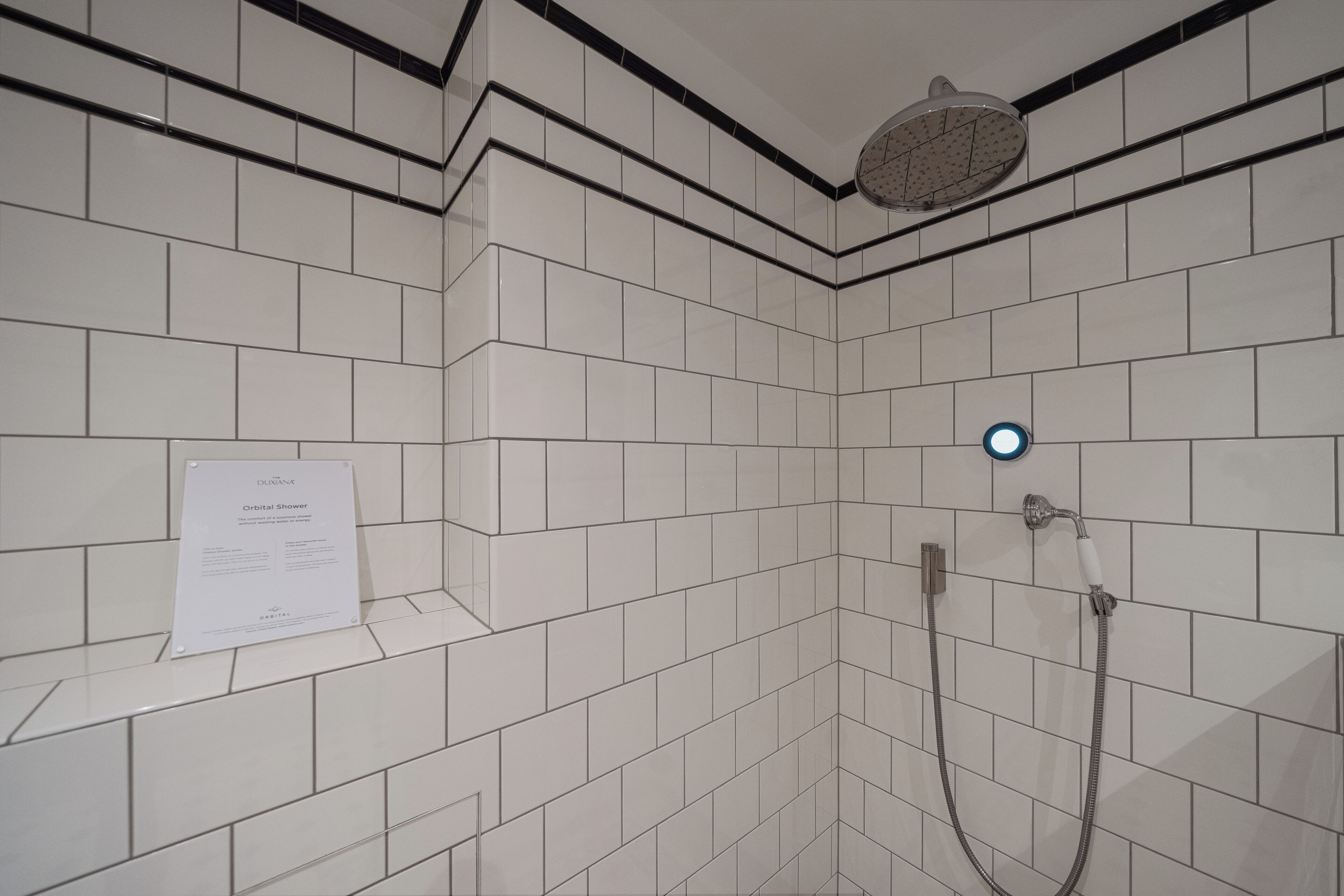 Ein Badezimmer mit weiß gefliesten Wänden, einer Duschbrause, einer Handbrause und einem Schild zur Erklärung der Orbitaldusche.