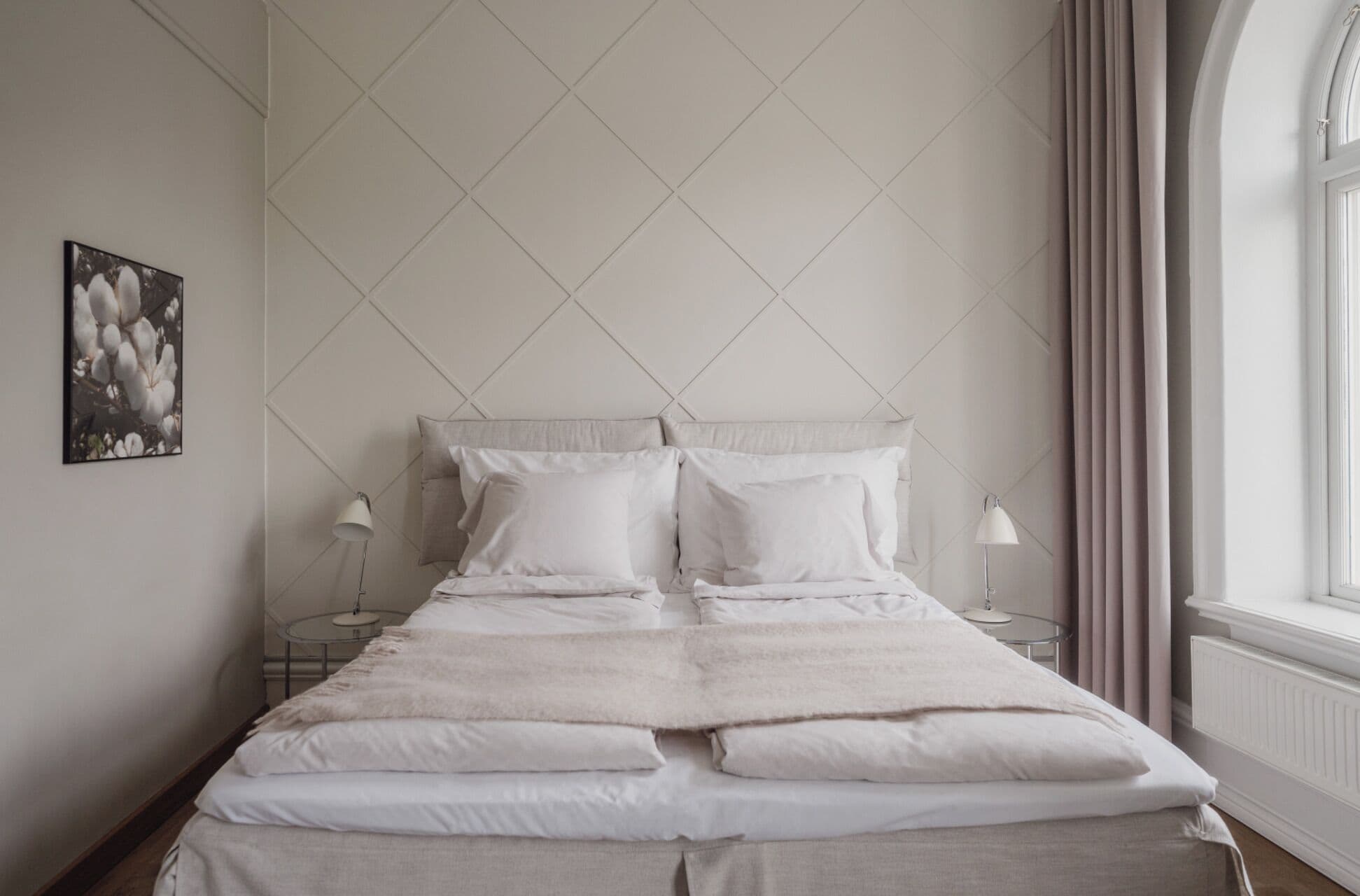 A neatly made bed with white linens and pillows, two bedside tables with lamps, a framed picture on the wall, and a window with curtains.