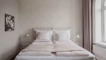 A neatly made bed with white linens and pillows, two bedside tables with lamps, a framed picture on the wall, and a window with curtains.