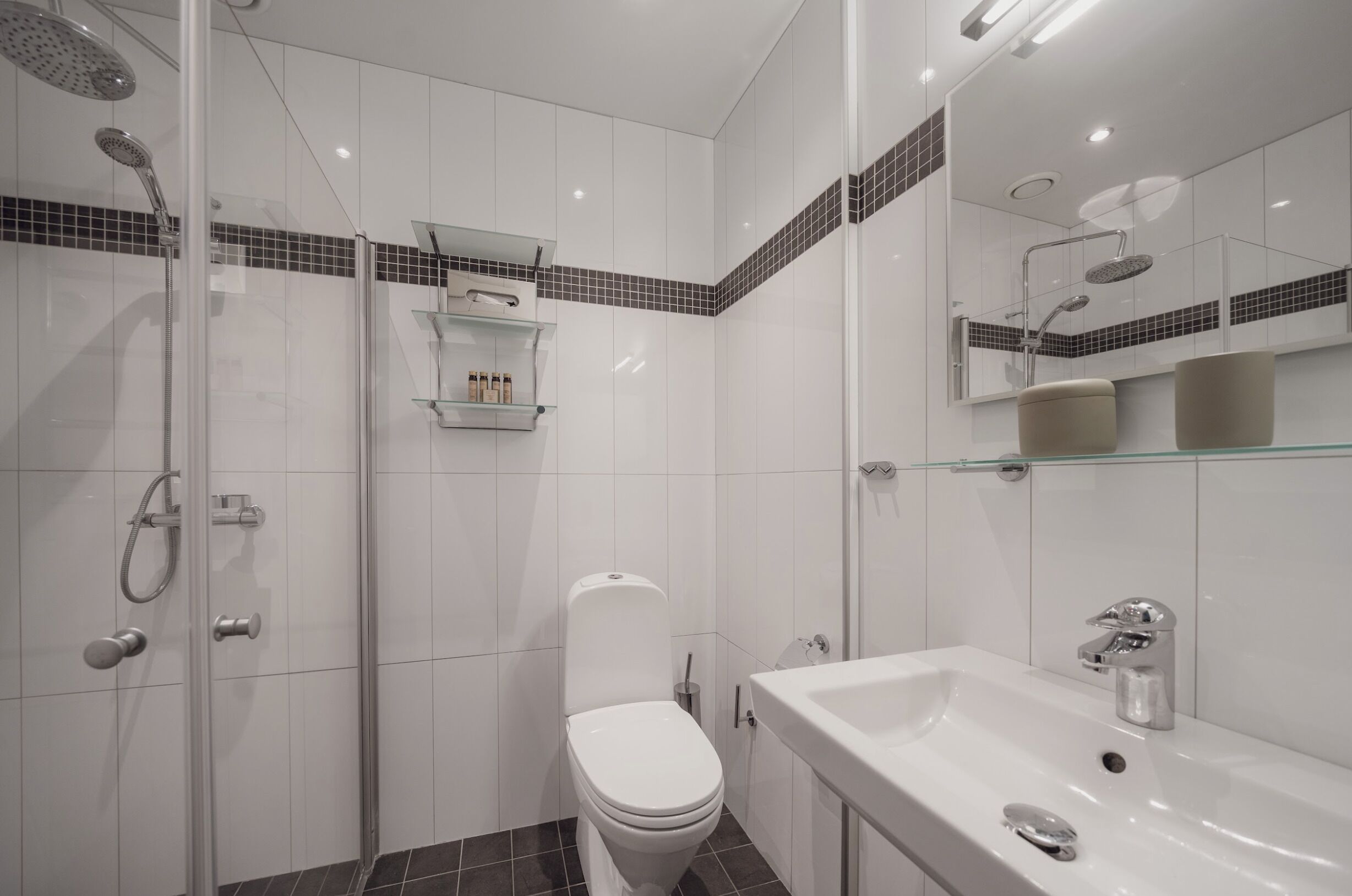 Un baño moderno con ducha de vidrio, inodoro blanco, repisa de vidrio con artículos de aseo y un espejo grande sobre el lavabo.