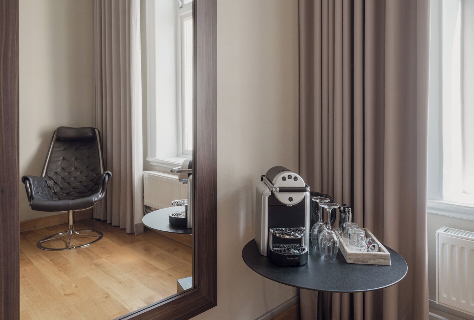 Ein Hotelzimmer mit einem schwarzen Ledersessel, einem kleinen runden Tisch mit Kaffeemaschine und Gläsern sowie einem Fenster mit Vorhängen.