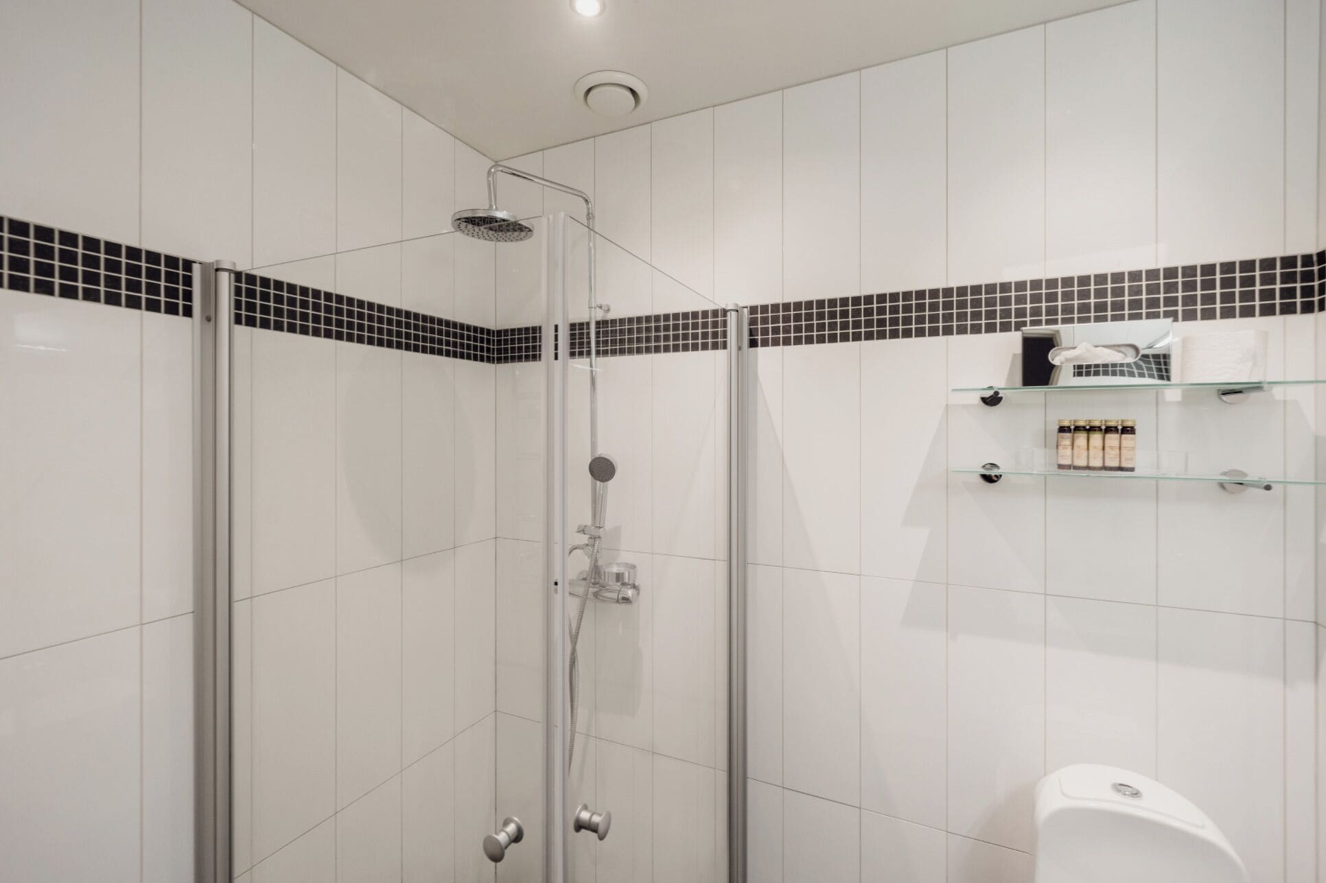 Un baño moderno con ducha envuelta en vidrio, azulejos blancos y un detalle decorativo con mosaicos negros.