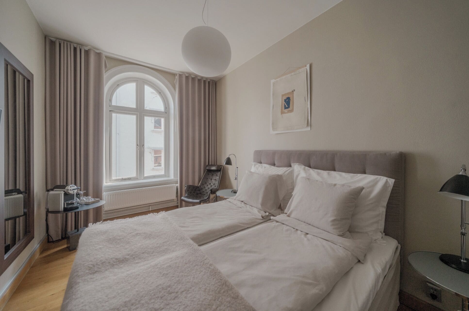 A hotel room with a large bed, a desk with a chair, a window with curtains, and a framed picture on the wall.