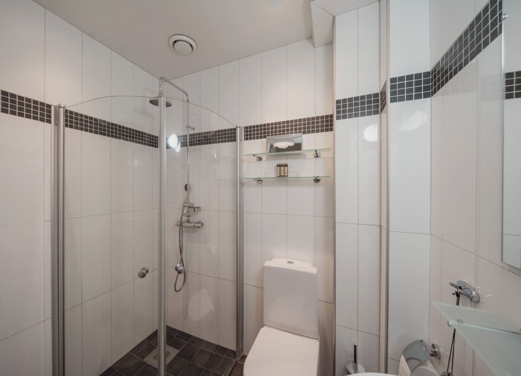Une salle de bain moderne avec une douche à parois de verre, des carreaux blancs et une petite étagère contenant des articles de toilette.